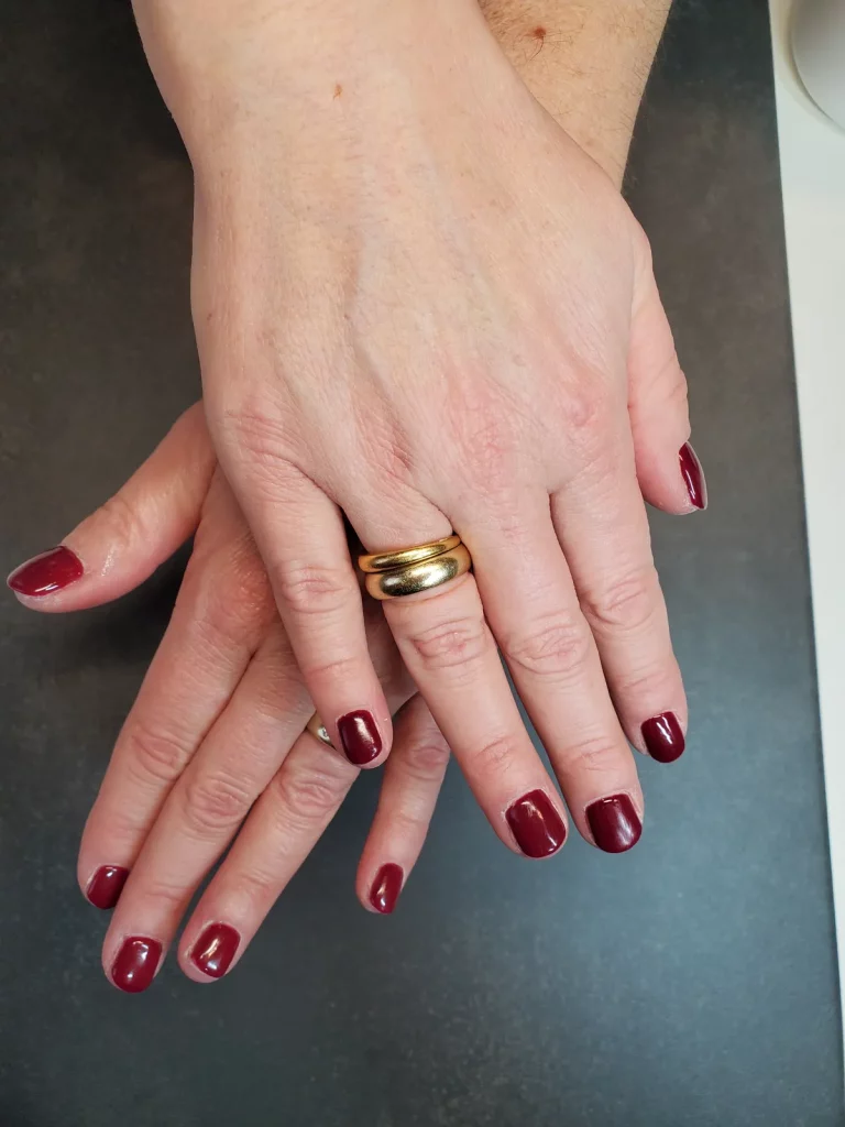 Hände übereinander mit glänzend bordeauxrotem Lack auf kurzen, sauber geformten Nägeln.