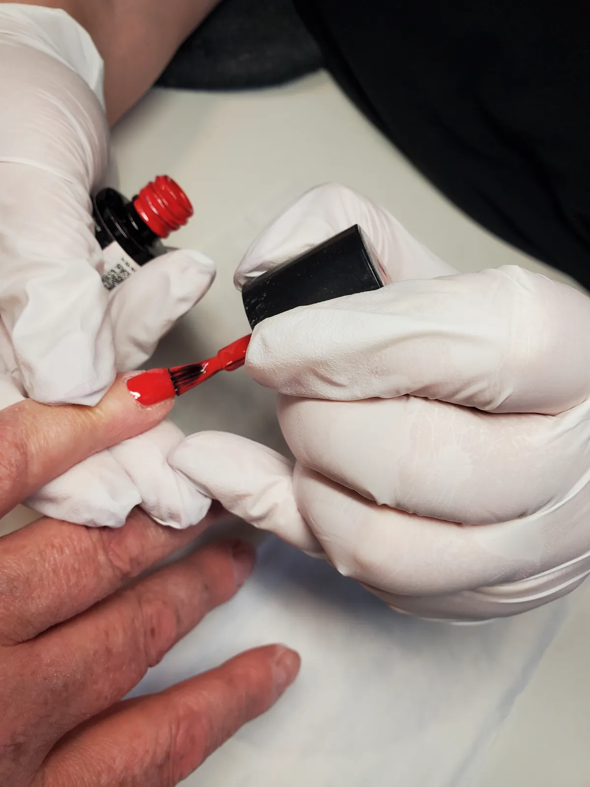 Roter Lack wird mit Pinsel auf einen Fingernagel aufgetragen, Nageldesignerin arbeitet mit Handschuhen.